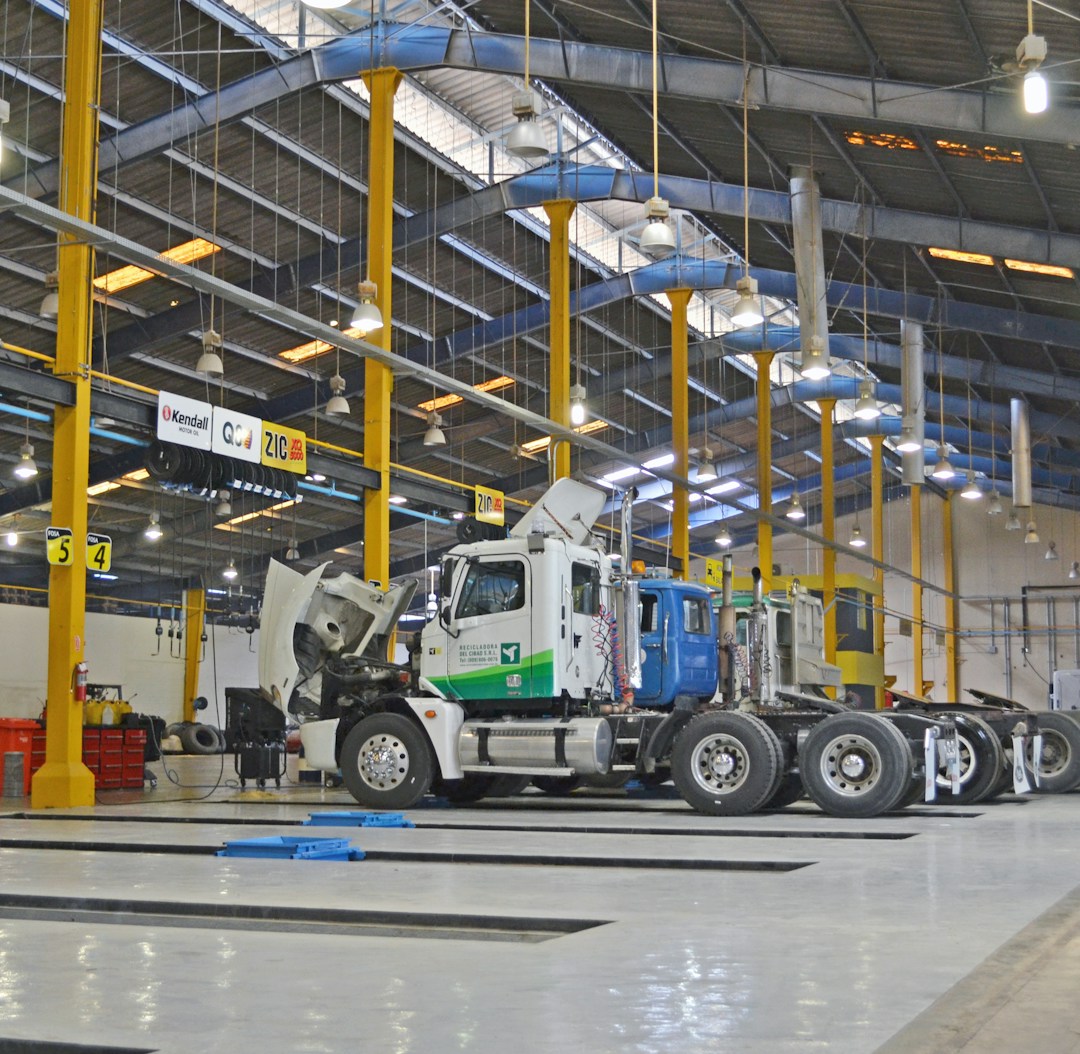 A large indoor vehicle maintenance facility with high ceilings and rows of overhead lights. Several trucks are parked in the space, with one having its hood open. Yellow support beams and signs hang from the ceiling. A person stands to the side, presumably working or supervising.