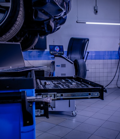 A car service or repair garage is depicted, with a vehicle hoisted on a lift. A laptop is placed on a nearby workstation with an open drawer filled with tools. The environment features tiled walls and floor, and equipment such as a tire changer and a balancing machine. Fluorescent lighting provides illumination.