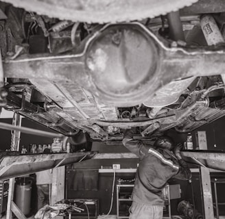 A mechanic is working underneath a vehicle that's elevated on a hydraulic lift. The scene captures various auto parts and tools scattered around the workshop. The environment appears to be a busy auto repair shop.