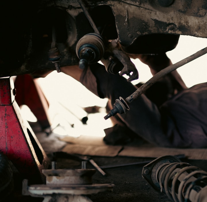 A mechanic is lying on the ground under a vehicle, working on its suspension system. The vehicle is supported by a red hydraulic jack stand. Various car parts and a tire are scattered nearby on the concrete floor, suggesting a repair or maintenance activity in a workshop environment.